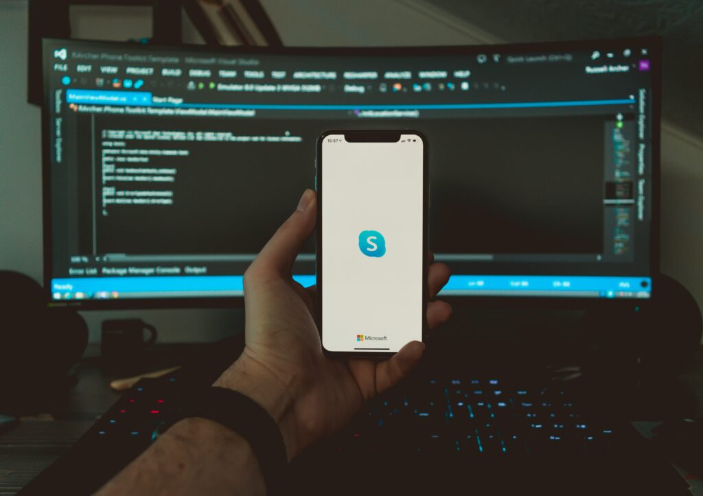 Person holding a phone with the Skype logo, in front of a computer with code on the monitor