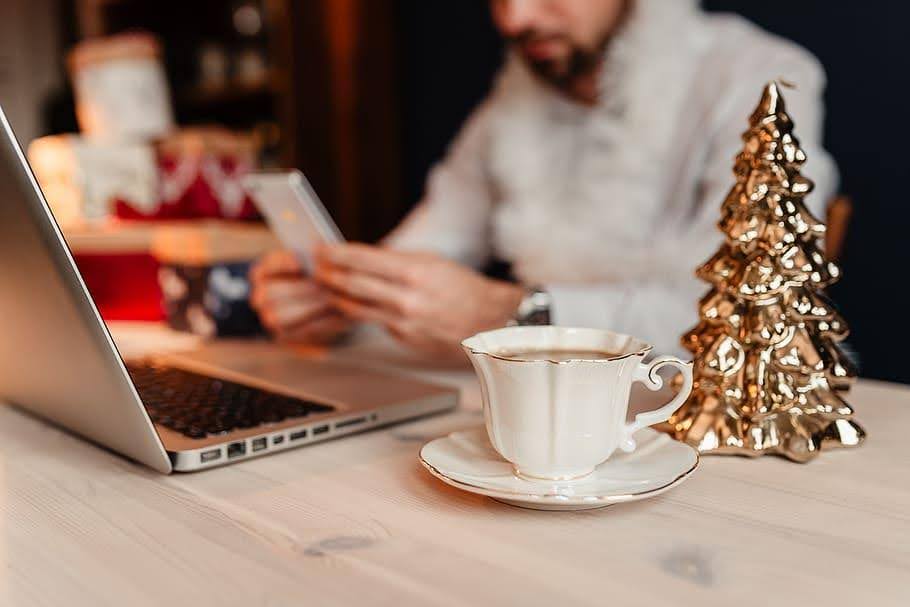 Man using phone by laptop with coffee cup and decorative gold Christmas tree on table