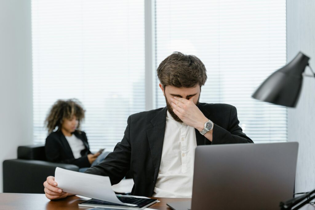 man seemingly stressed shuts his eyes closed in front of papers and laptop