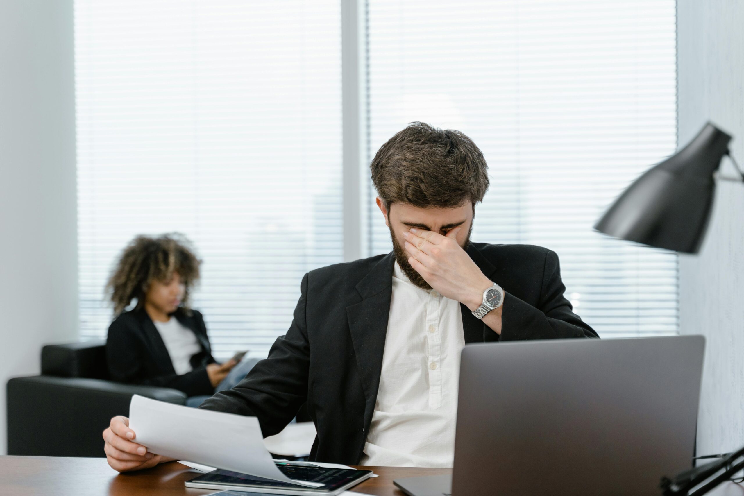 man seemingly stressed shuts his eyes closed in front of papers and laptop
