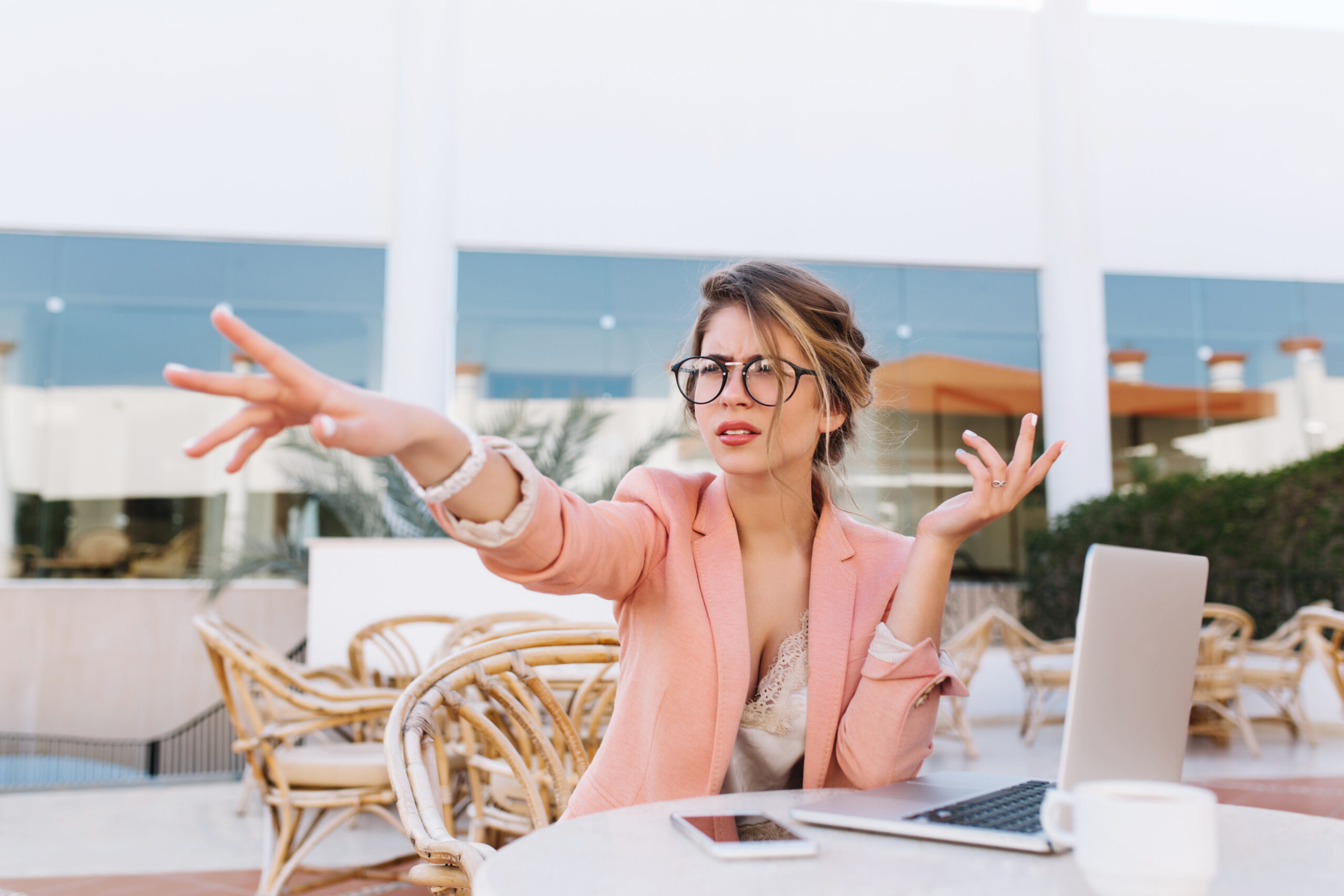 Young business woman sitting in outdoor cafe with laptop on table, serious lady pointing with hand direction, saw something in afait. Wearing stylish pink jacket, glasses, white watches..